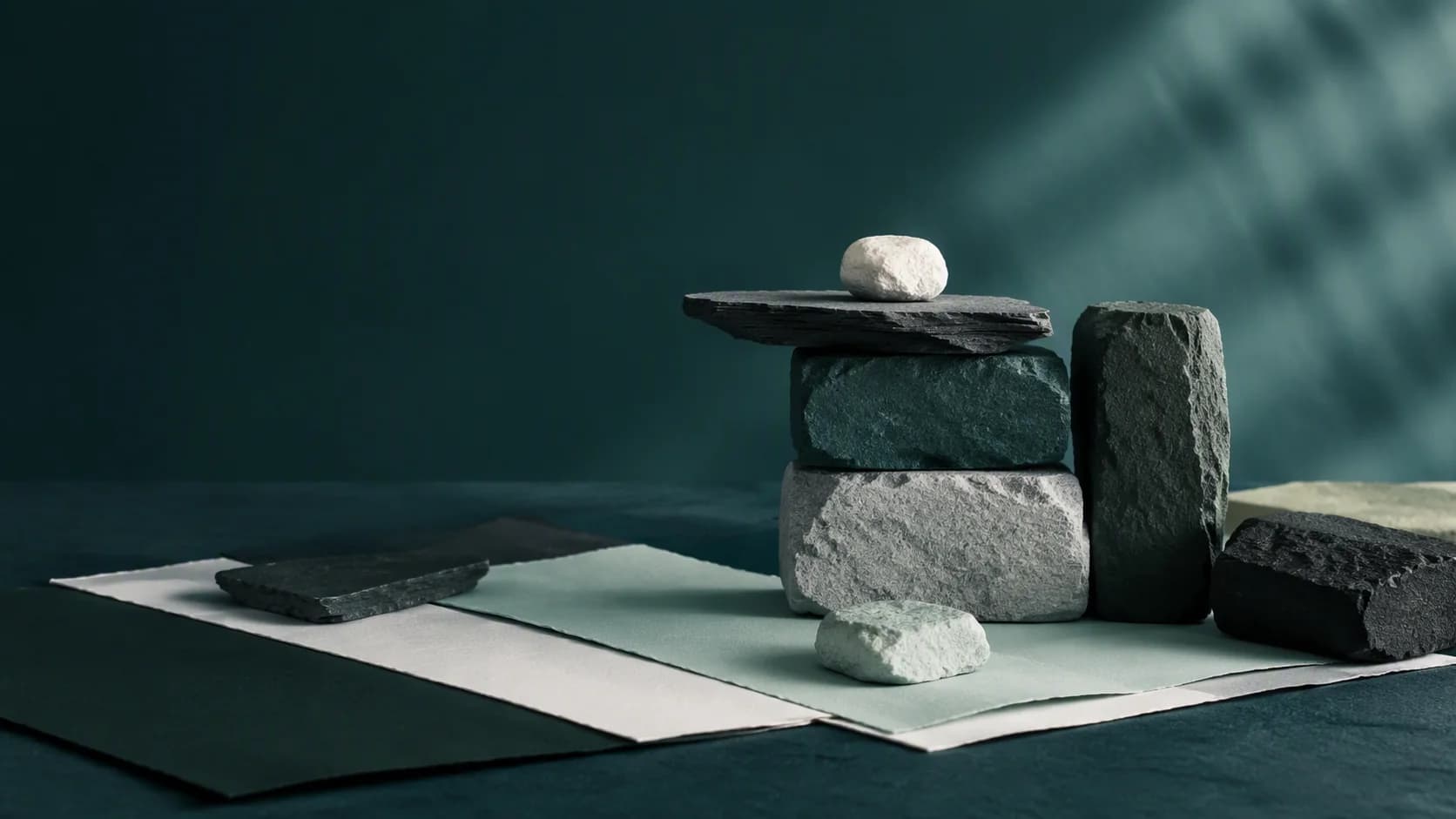 An abstract arrangement of balanced stone blocks and layered teal paper-like planes in a quiet landscape.